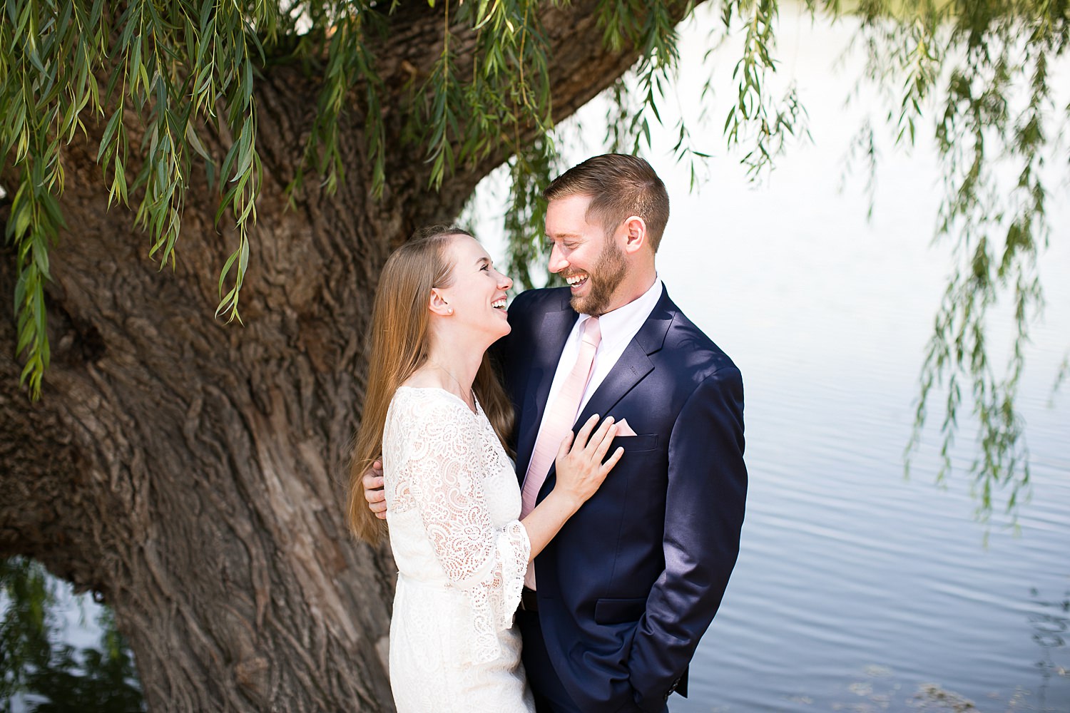 Couple in wedding attire laughs under willow tree at Chicago Botanic Garden.